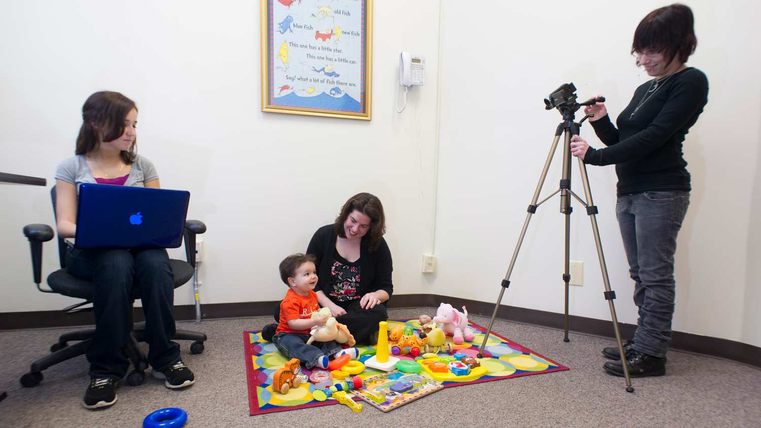 Two people recording a baby playing on a play mat with another adult